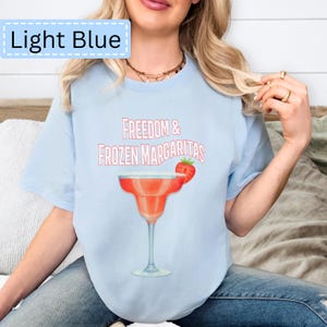 May include: Light blue t-shirt with the text "Freedom & Frozen Margaritas" above a watercolor illustration of a margarita cocktail with a strawberry garnish. The words "Light Blue" are in a light blue box in the upper left corner.