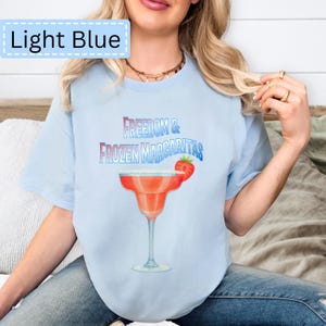 May include: Light blue t-shirt with the text "Freedom & Frozen Margaritas" and a margarita glass graphic. The margarita glass is red with a white rim and a strawberry garnish. The shirt is a light blue color.