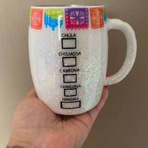 May include: White ceramic mug with a glittery finish and colorful paper banners around the top. The mug has five black boxes with the words "Chula", "Chismosa", "Cabrona", "Chingona", and "Perrona" printed on them.