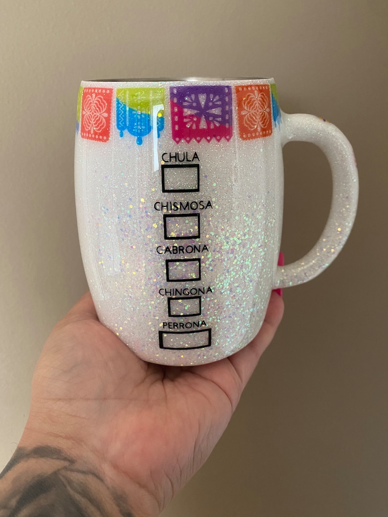 May include: White ceramic mug with a glittery finish and colorful paper banners around the top. The mug has five black boxes with the words "Chula", "Chismosa", "Cabrona", "Chingona", and "Perrona" printed on them.