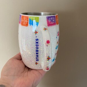 May include: White stainless steel tumbler with a glittery finish and a colorful Mexican-inspired design. The tumbler features the Starbucks logo and a handle.