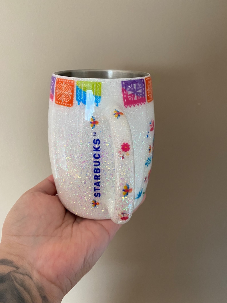 May include: White stainless steel tumbler with a glittery finish and a colorful Mexican-inspired design. The tumbler features the Starbucks logo and a handle.