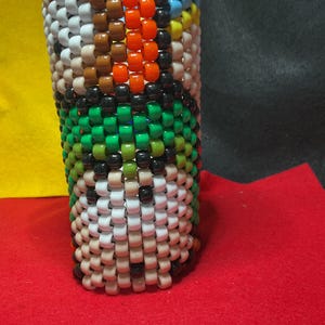 May include: A cylindrical object made from colourful beads. The design features white, brown, orange, black, green, and blue beads. The beadwork is tightly woven, creating a textured surface. The object is set against a red surface.