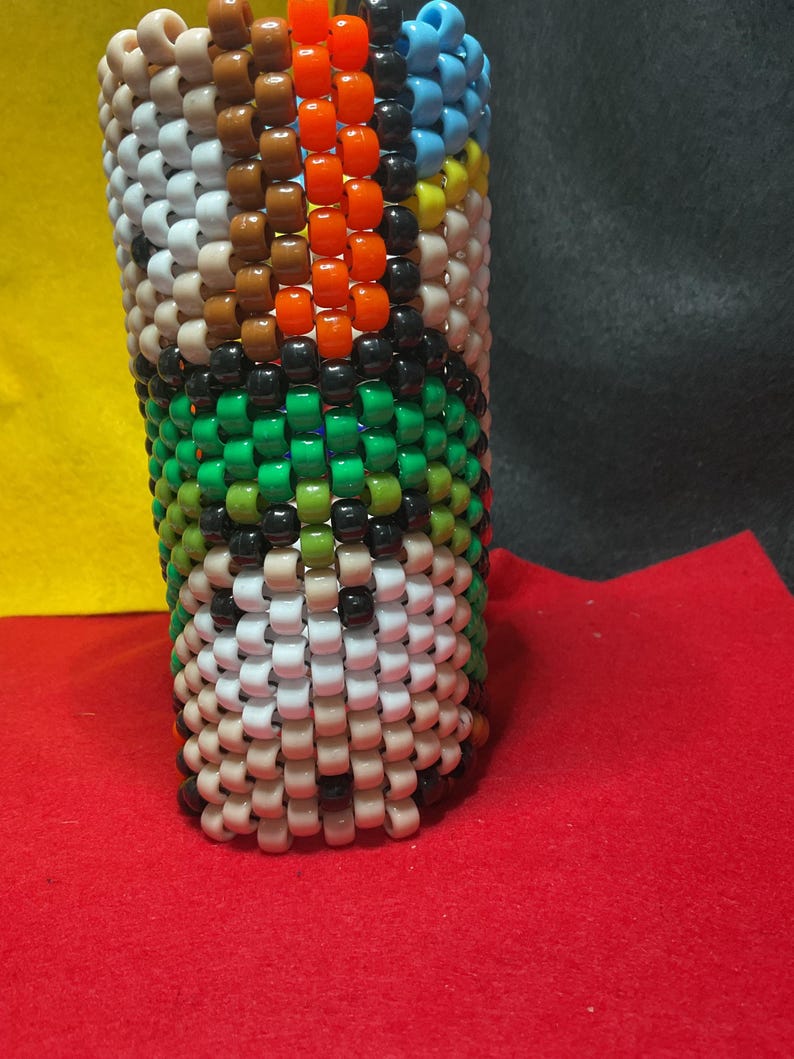 May include: A cylindrical object made from colourful beads. The design features white, brown, orange, black, green, and blue beads. The beadwork is tightly woven, creating a textured surface. The object is set against a red surface.