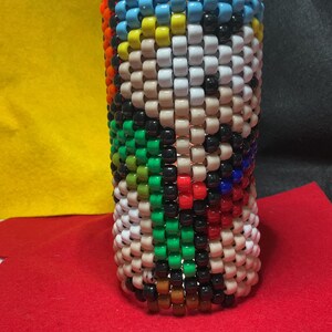 May include: A bottle cover made from colourful beads, featuring a pattern of white, tan, black, green, red, blue, yellow, and orange beads. The top edge is lined with light blue beads. The design is cylindrical.