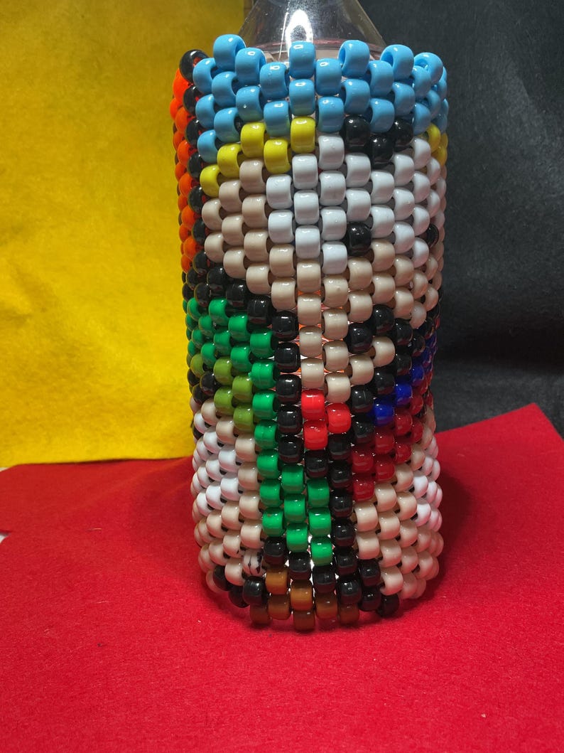 May include: A bottle cover made from colourful beads, featuring a pattern of white, tan, black, green, red, blue, yellow, and orange beads. The top edge is lined with light blue beads. The design is cylindrical.