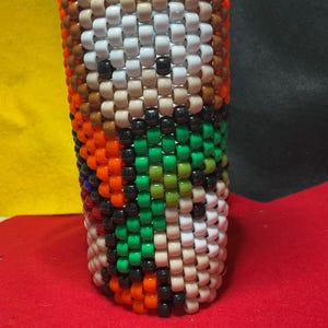 May include: A cylindrical object made of beads in a variety of colours, including white, black, orange, green, and brown. The beads are arranged in a pattern, creating a textured surface. The object is standing upright.