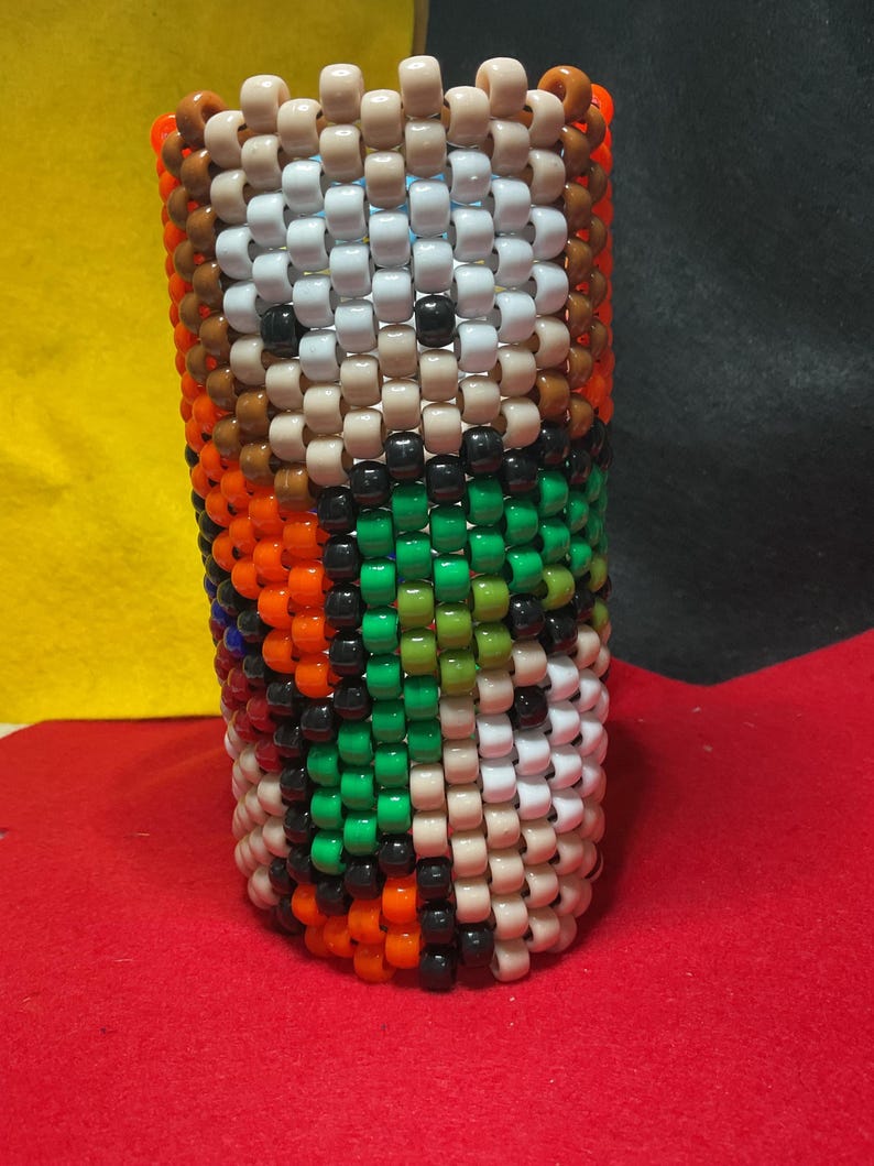 May include: A cylindrical object made of beads in a variety of colours, including white, black, orange, green, and brown. The beads are arranged in a pattern, creating a textured surface. The object is standing upright.