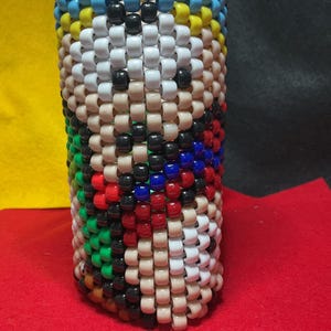 May include: A cylindrical container made of beads, featuring a design in blue, white, tan, black, red, green, and yellow. The top edge is a row of blue beads. The item is made of small, colourful beads.