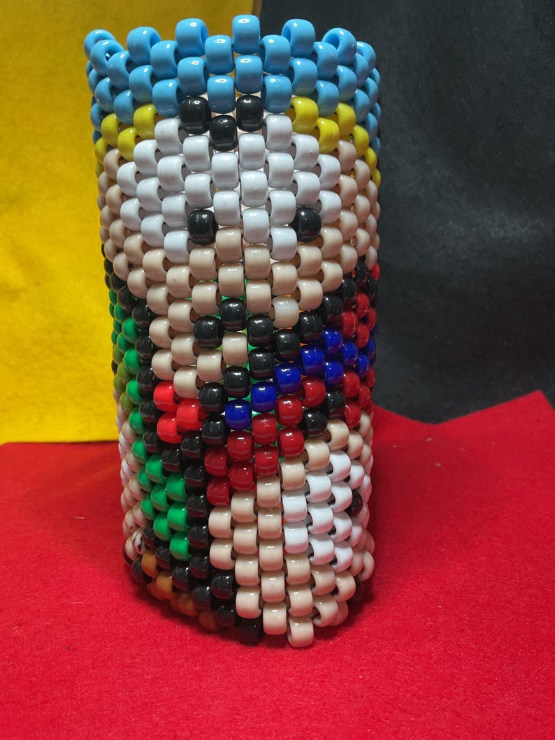 May include: A cylindrical container made of beads, featuring a design in blue, white, tan, black, red, green, and yellow. The top edge is a row of blue beads. The item is made of small, colourful beads.