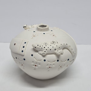 May include: A white ceramic vase with a round shape, featuring two sculpted lizards with black spots. The vase is decorated with blue and red floral patterns and a rectangular accent. The vase has a small opening at the top.