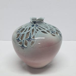 May include: A ceramic vase with a rounded body featuring a gradient from soft pink at the base to light blue at the top. The upper section has an intricate, open-work lattice design in dark blue and grey, resembling stylized floral petals.