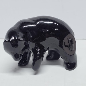 May include: A glossy black ceramic bear figurine in a walking pose. The bear features a circular matte gray patch on its rear flank, which is embossed with a dark, stylized paw print design. The surface of the bear is smooth and reflective.