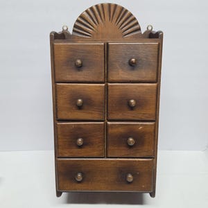 May include: A small, vintage wooden cabinet with eight drawers and a decorative sunburst design at the top. The cabinet is made of dark brown wood and features small, round knobs on each drawer. It has a rustic, antique aesthetic.