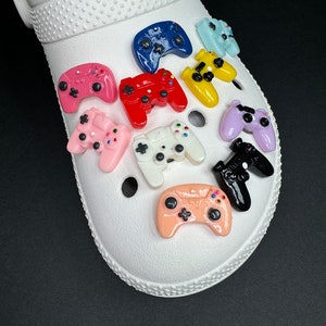 May include: A white shoe with a variety of colorful game controller charms attached to it. The charms are in various colors, including pink, red, blue, yellow, and black.