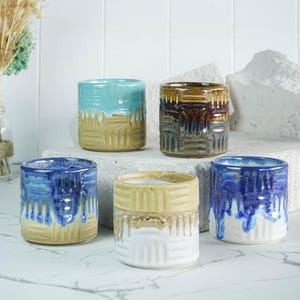 May include: Five handmade ceramic cups in shades of blue, turquoise, brown, and yellow. Each cup has a unique glaze and textured design. The cups are cylindrical and appear to be small, perfect for espresso or small beverages.