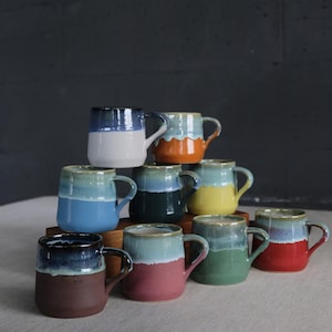 May include: A collection of handmade ceramic mugs in various colors, including blue, orange, yellow, green, and red. Each mug features a two-tone design with a contrasting color on the bottom half and a glazed top. The mugs are stacked on a wooden surface.