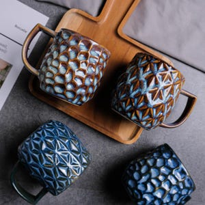 Könnte beinhalten: Vier Keramikbecher mit strukturierten Oberflächen und Henkeln. Die Becher haben blaue und braune Farbtöne mit geometrischen und Wabenmustern. Sie sind auf einem Holzserviertablett angeordnet, vor einem grauen Hintergrund.