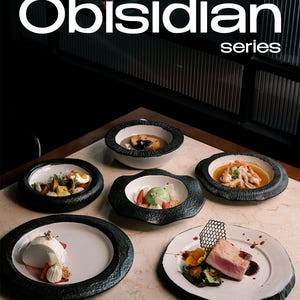 Puede incluir: Una colección de platos de la "serie Obisidian" se exhibe sobre una superficie de mármol. Los platos y cuencos tienen un interior blanco y un borde negro texturizado. Se presentan varias creaciones culinarias, incluyendo postres y platos salados.