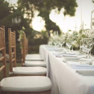Puede incluir: Una mesa de comedor larga cubierta con un mantel blanco, dispuesta con cristalería transparente, platos blancos y velas cónicas blancas altas. Una fila de sillas de madera con asientos acolchados blancos está dispuesta a lo largo.