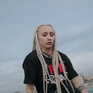 May include: A person wearing a black t-shirt with red and white text and long blonde braids. The person is wearing a silver chain and bracelets. The background is a cloudy sky and industrial buildings.