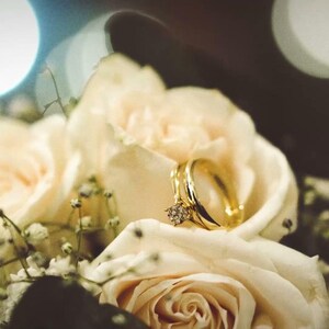 Puede incluir: Un anillo de oro con un diamante de talla redonda engastado en el centro, descansando sobre un grupo de rosas de color crema pálido y pequeñas flores blancas de paniculata. El aro de metal está pulido y refleja la luz sobre los pétalos.