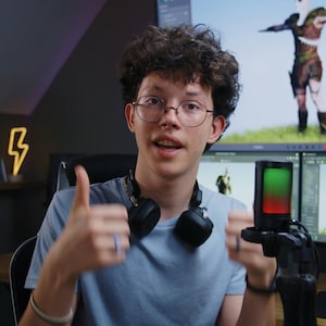 May include: A young person with curly hair and glasses gives a thumbs-up gesture. They are wearing a light blue t-shirt and headphones. Two computer monitors display a 3D model of a character. A neon lightning bolt decorates the wall.