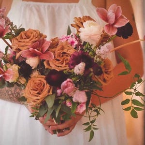 Puede incluir: Un ramo de flores que presenta una mezcla de rosas color óxido, flores de color púrpura intenso, tulipanes rosa suave y ranúnculos blancos, acentuado con follaje verde y tallos pequeños texturizados, reunidos en un arreglo de flores frescas.