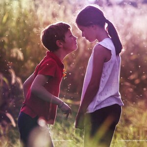 以下が含まれることがあります： 赤いポロシャツとダークカラーのパンツを着用した子供が、白いノースリーブのトップスとダークカラーのパンツを着用した別の子供と向かい合っています。夕暮れ時の背の高い草が生い茂る野原に立ち、柔らかな光と空中に浮かぶ粒子が見えます。