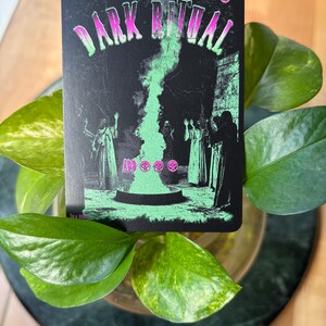 Pode incluir: Um cart&atilde;o preto com as palavras "DARK RITUAL" em rosa e verde. O cart&atilde;o apresenta uma chama verde e figuras em vestes. O cart&atilde;o &eacute; cercado por folhas verdes.
