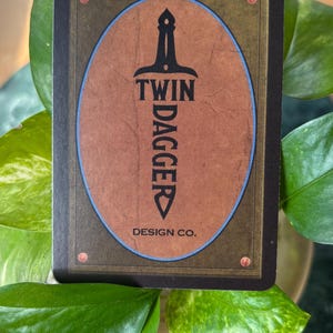 Pode incluir: Um cart&atilde;o retangular com um design oval marrom apresentando uma adaga preta e as palavras "TWIN DAGGER" e "DESIGN CO." em branco. O cart&atilde;o tem uma borda escura com rebites cor de cobre. O cart&atilde;o &eacute; cercado por folhas verdes.