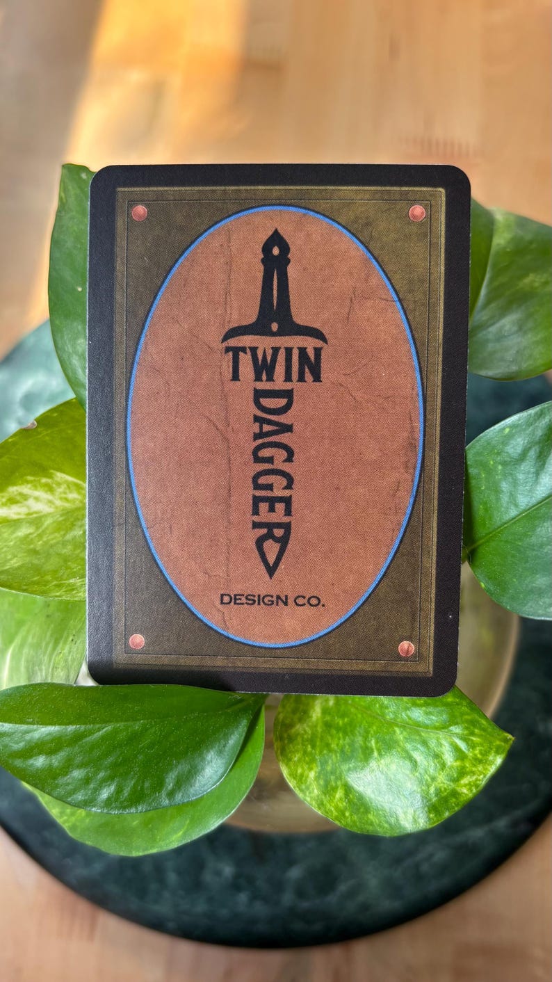 Pode incluir: Um cart&atilde;o retangular com um design oval marrom apresentando uma adaga preta e as palavras "TWIN DAGGER" e "DESIGN CO." em branco. O cart&atilde;o tem uma borda escura com rebites cor de cobre. O cart&atilde;o &eacute; cercado por folhas verdes.