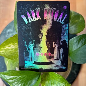 Pode incluir: Um cart&atilde;o preto com as palavras "DARK RITUAL" em letras iridescentes. A imagem mostra figuras em vestes em volta de um caldeir&atilde;o com uma chama colorida. O cart&atilde;o tem um acabamento hologr&aacute;fico.