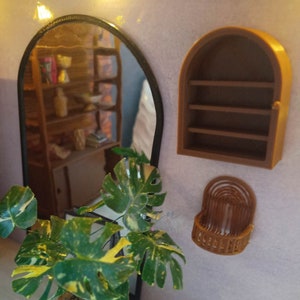 May include: A miniature dollhouse room with a black oval mirror, a brown arched shelf, a brown wicker wall hanging, and a green and yellow potted plant.