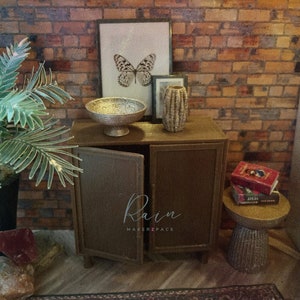 May include: A brown wooden cabinet with two doors, a silver bowl, a gold vase, and a framed print of a butterfly. The cabinet is on a brick wall background. The cabinet has the text "Rain Makerzpace" on the front.