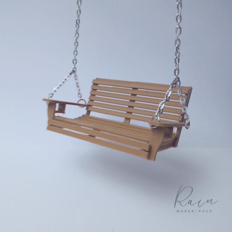 May include: A miniature wooden swing with a slatted seat and back, suspended by silver-colored chains. The swing is a light brown color and appears to be a handcrafted item. The image is set against a plain white background.