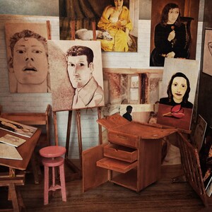 Puede incluir: Un estudio de artista en miniatura con pinturas de retratos y figuras. El estudio cuenta con una mesa de dibujo de madera, un taburete rosa y un caballete de madera con un retrato. Varias pinturas se exhiben en las paredes.