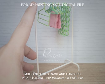 May include: A white metal clothes rack with green hangers and a pink garment. The rack is a 1:12 miniature model inspired by IKEA's MULIG clothes rack. The text "FOR 3D PRINTING - STL DIGITAL FILE" and "Rain MAKERSPACE" are visible in the image. The text "MULIG CLOTHES RACK AND HANGERS" and "IKEA - Inspired 1:12 Miniature - 3D STL File" are also visible in the image.