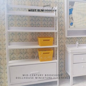 Pode incluir: Estante branca com uma gaveta e duas cestas de armazenamento amarelas. A estante &eacute; inspirada nos m&oacute;veis da West Elm e &eacute; uma miniatura de casa de bonecas em escala 1:12. O texto "MID-CENTURY BOOKSHELF DOLLHOUSE MINIATURE 1:12 SCALE" est&aacute; impresso abaixo da estante.