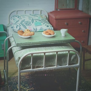 Puede incluir: Una cama de hospital de metal blanco con un colchón verde y una almohada con estampado floral. Una mesa pequeña con una jarra blanca y dos platos de pasteles está al lado de la cama.