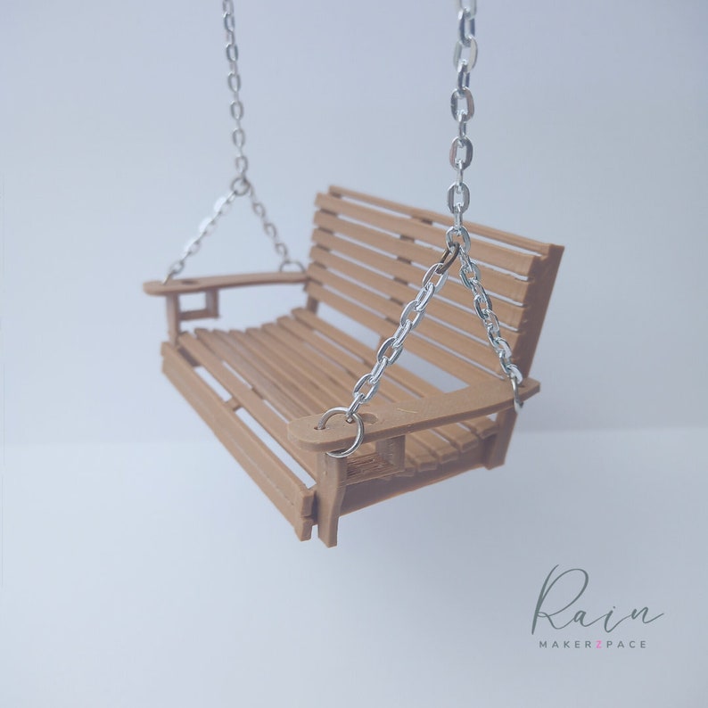 May include: A miniature wooden swing with a slatted seat and back, suspended by silver chains. The swing is a light brown color and has armrests. The background is a soft, blurred white. The logo "Rain MAKERZPACE" is in the bottom right corner.
