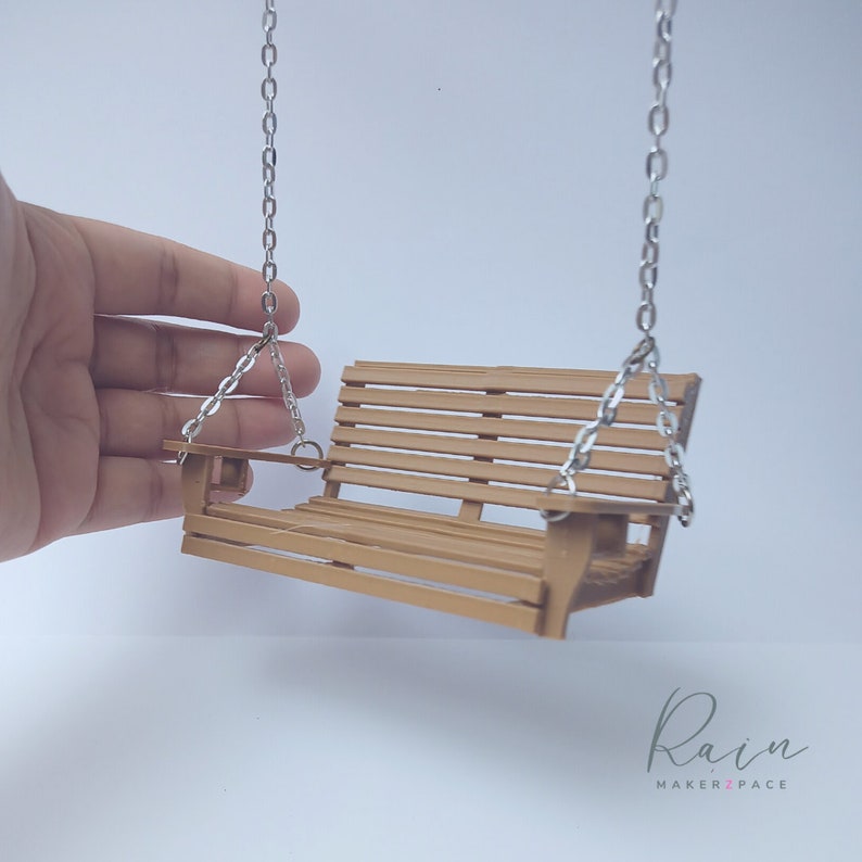 May include: A miniature wooden swing with a slatted seat and back, suspended by silver-colored chains. The swing is a warm brown color and appears to be a handcrafted item. The background is plain white.