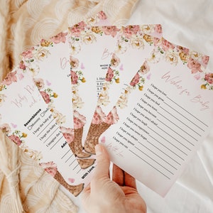 Op de afbeelding: Een set bedrukte papieren kaarten voor een babyshower met een bloemenrand van roze en cr&egrave;mekleurige bloemen en kleine vlinders. De kaarten bevatten tekst zoals Wishes for Baby, Baby Resemblance en lijnen om antwoorden in zwarte inkt te schrijven.
