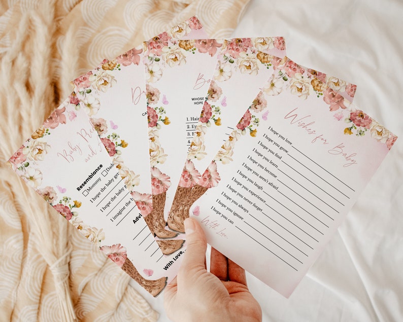 Op de afbeelding: Een set bedrukte papieren kaarten voor een babyshower met een bloemenrand van roze en cr&egrave;mekleurige bloemen en kleine vlinders. De kaarten bevatten tekst zoals Wishes for Baby, Baby Resemblance en lijnen om antwoorden in zwarte inkt te schrijven.