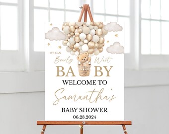 May include: A rectangular white sign featuring a hot air balloon made of gold, beige, and white balloons carrying a teddy bear in a basket. The text reads: WE CAN Bearly Wait BABY WELCOME TO Samantha's BABY SHOWER 06.28.2024, with gold stars and clouds.