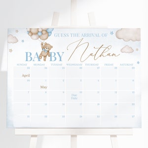 Op de afbeelding: Een blauwe en witte baby shower-kalender met een teddybeer die ballonnen vasthoudt. De kalender heeft de titel "Raad de aankomst van Nathan" en bevat de maanden april en mei.
