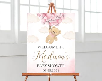 May include: A rectangular sign featuring a tan teddy bear holding a cluster of pink balloons against a white background with soft pink clouds. The text reads: WELCOME TO Madison's BABY SHOWER 03.23.2024. The sign is displayed on a wooden easel.