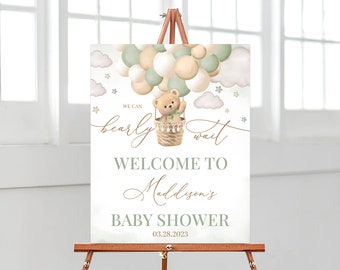 May include: A welcome sign for a baby shower with a teddy bear in a hot air balloon made of green and beige balloons. The sign says "We can bearly wait" and "Welcome to Maddison's Baby Shower 03.28.2023".