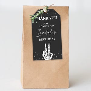May include: A brown paper gift bag with a black tag that reads "THANK YOU FOR COMING TO Isabel's BIRTHDAY". The tag features a white skeleton hand making a peace sign. A sprig of greenery is attached.