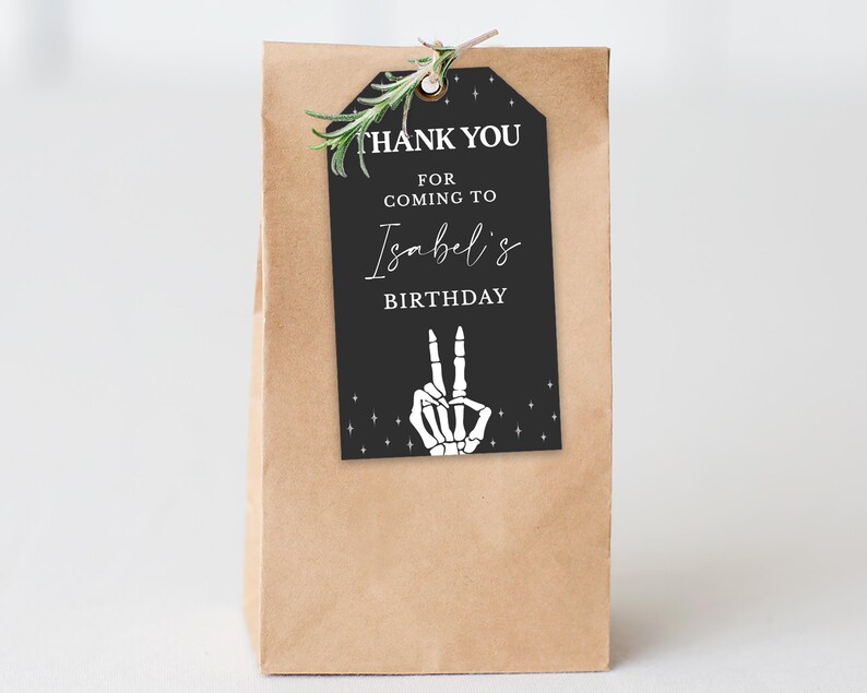 May include: A brown paper gift bag with a black tag that reads "THANK YOU FOR COMING TO Isabel's BIRTHDAY". The tag features a white skeleton hand making a peace sign. A sprig of greenery is attached.